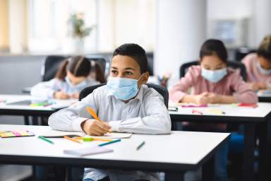 Boy in mask sitting at desk in classroom