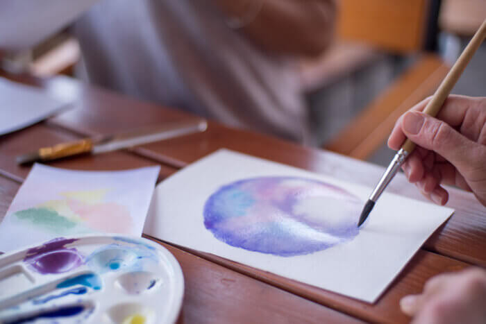 Closeup of hands painting an image of a purple, blue and pink circle with a watercolor brush and paint.
