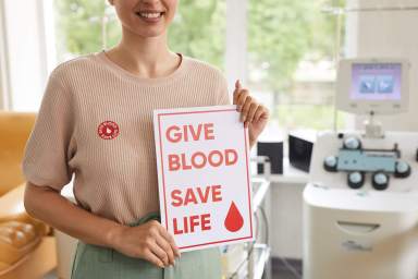 Close-up of young woman smiling and holding a poster that reads "Give Blood / Save Life." Take part in a blood drive at Empire City Casino.